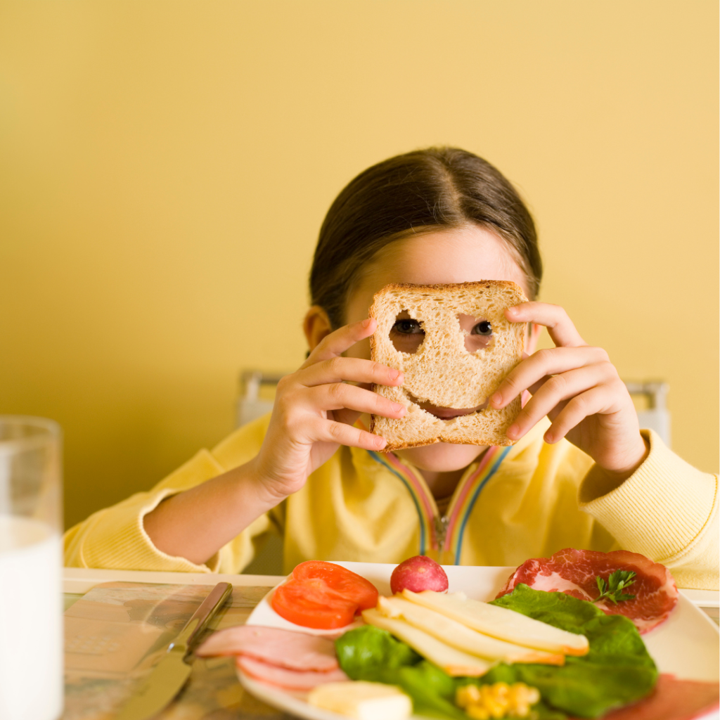 child playing with food, holding a piece of bread that has a smiley face cut out of it up to their face